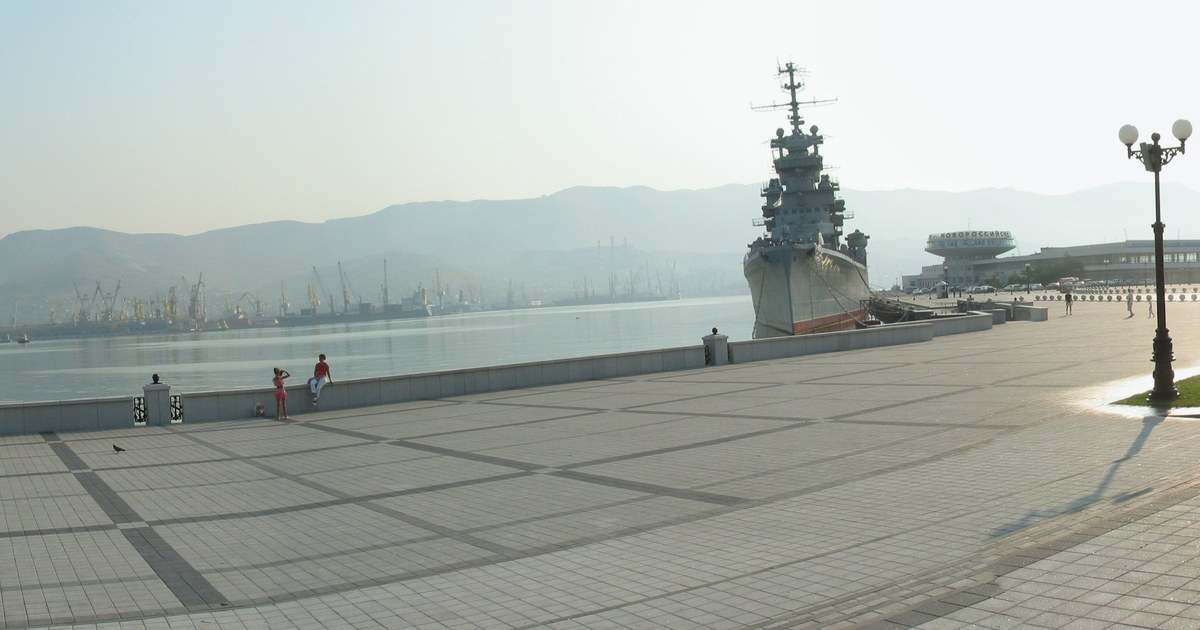 Panoramic view of Novorossiysk harbour with port cranes, waterfront infrastructure, mountains and a naval vessel.