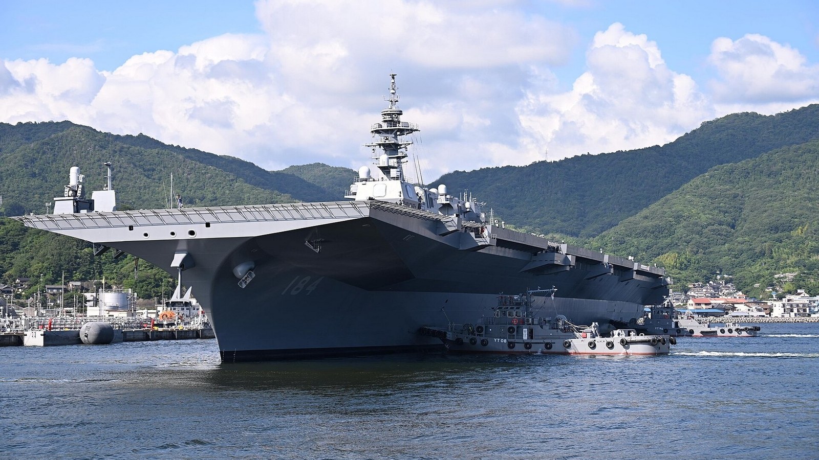 JS Kaga, hull number 184, returns to Kure Naval Base with wooded hills behind it.