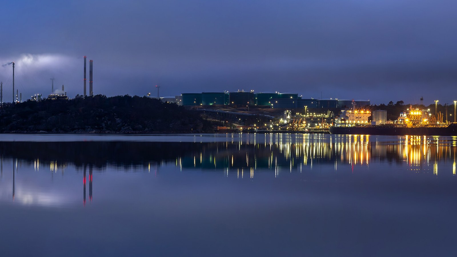 Storage tanks and processing units at an oil refinery are lit at dusk beside a waterway.