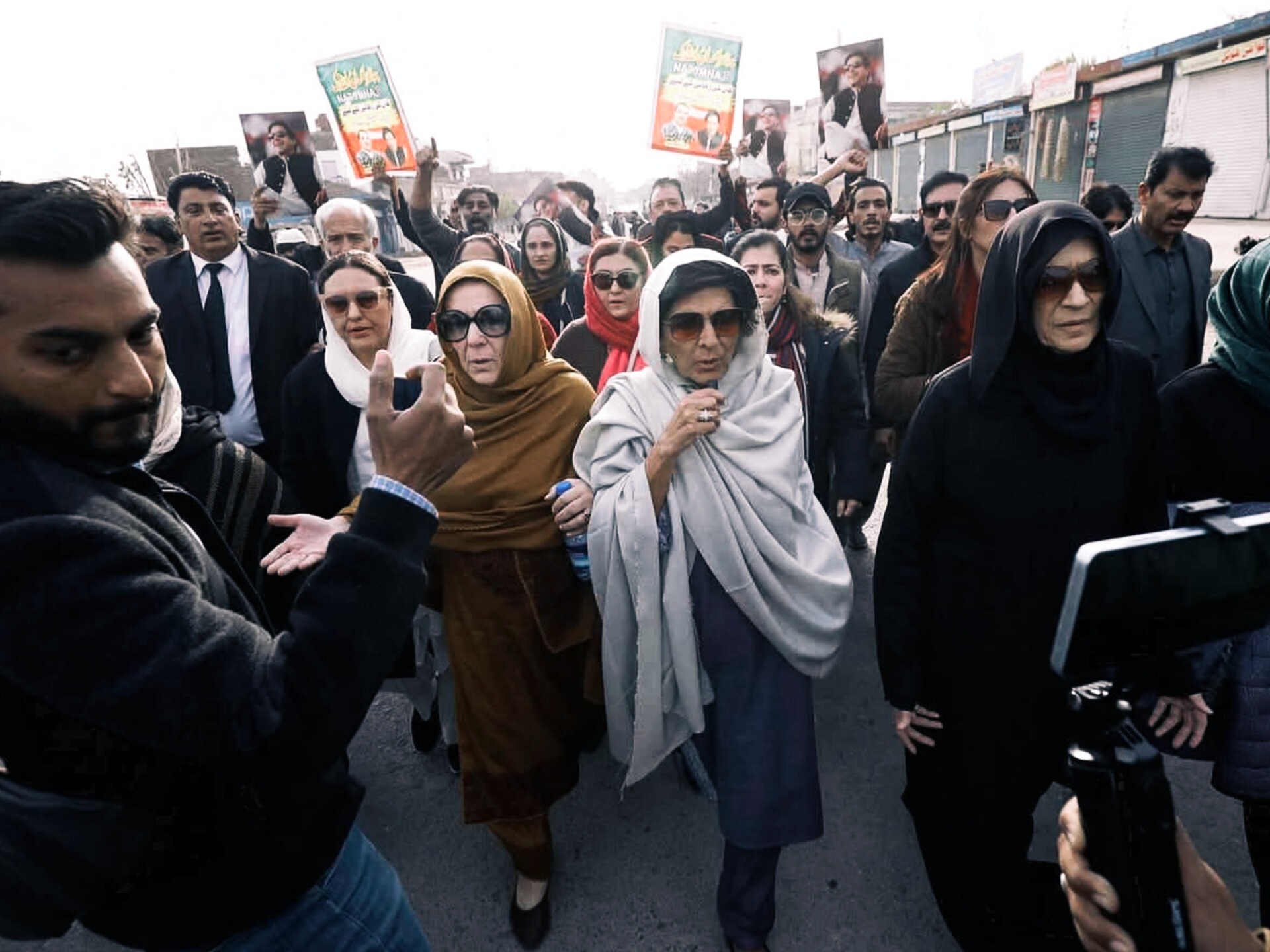 A crowd walks through a street during a political gathering in Pakistan, with several people holding portraits and party placards.