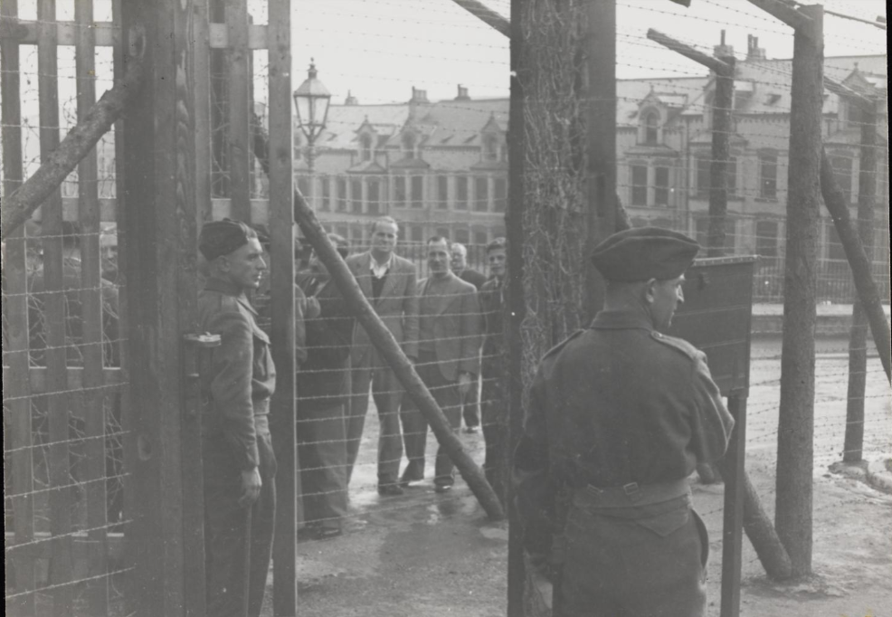 Photograph of internees and guards alongside the wire fence surrounding Hutchinson Internment Camp