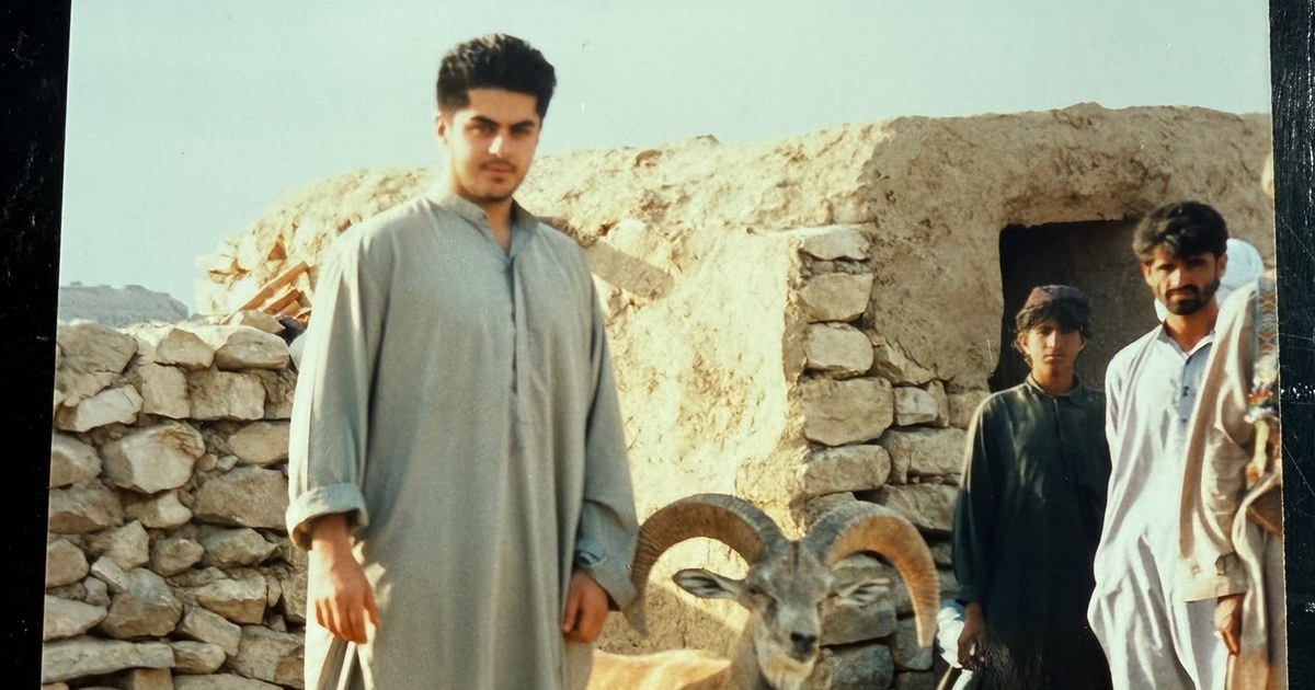 A younger Mehran Marri Baloch standing beside a ram in Kabul, with several people and stone buildings behind him.