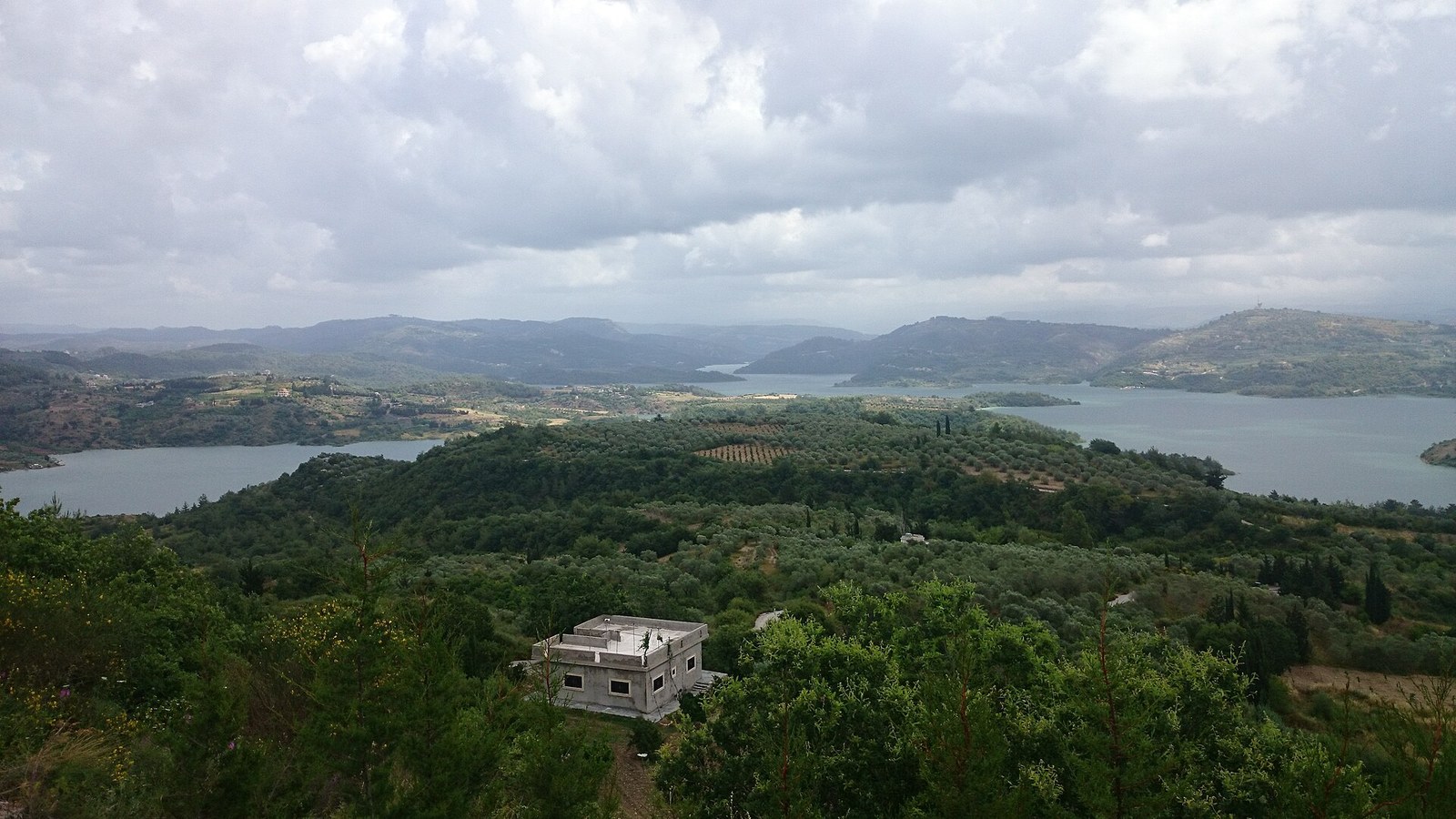 A wide view of green hills, a lake and scattered buildings in the Latakia countryside.