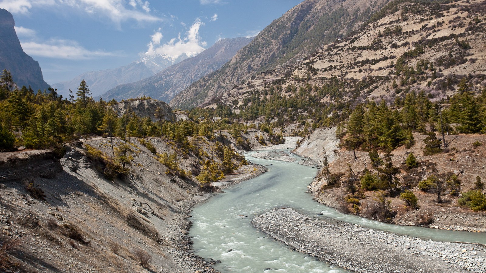 A turquoise glacial river winds through a steep Himalayan valley beneath distant snow-capped peaks.